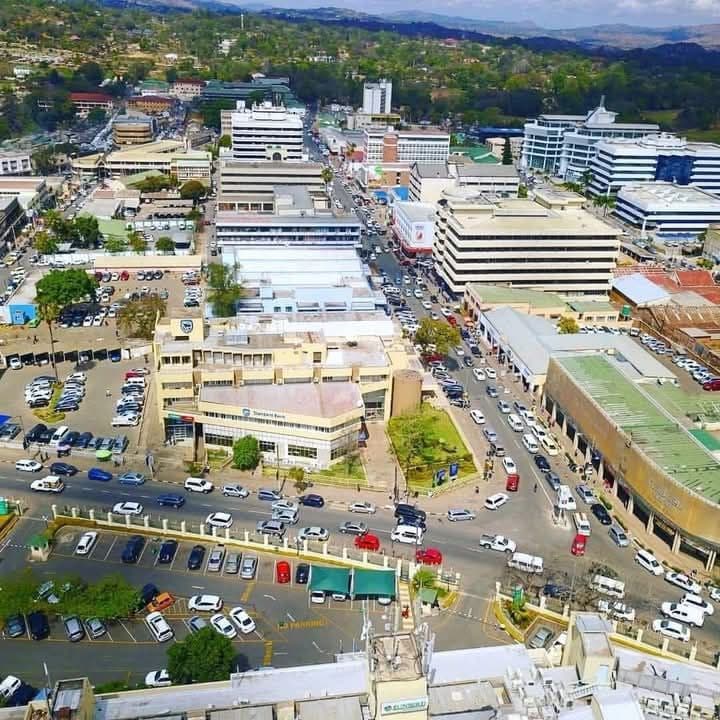 Aerial view of a busy city center with multiple office buildings, cars parked in lots, and traffic on the streets, surrounded by green hills in the distance.