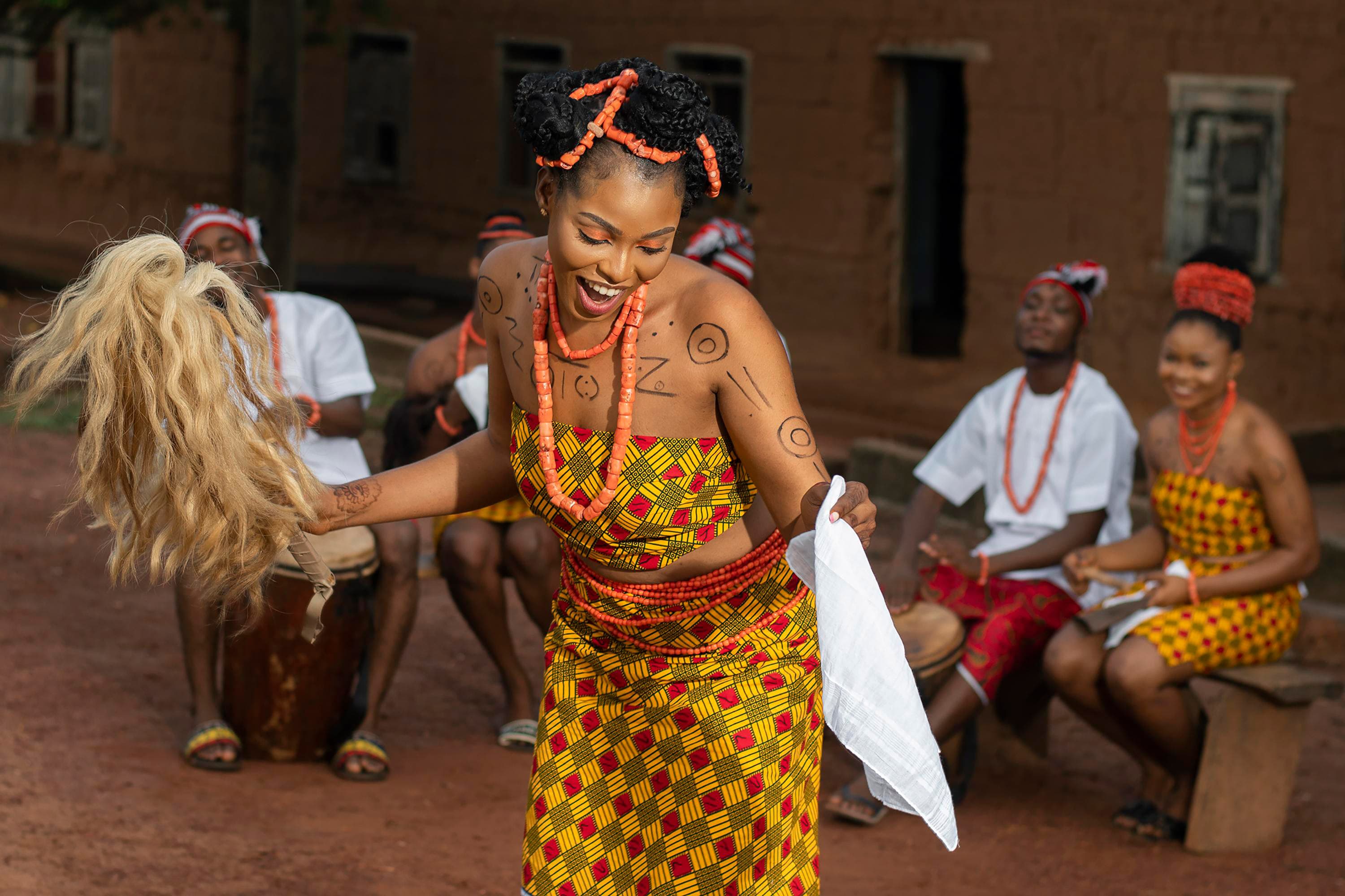 Smiling woman in traditional African attire performing a cultural dance with drummers and singers in the background.